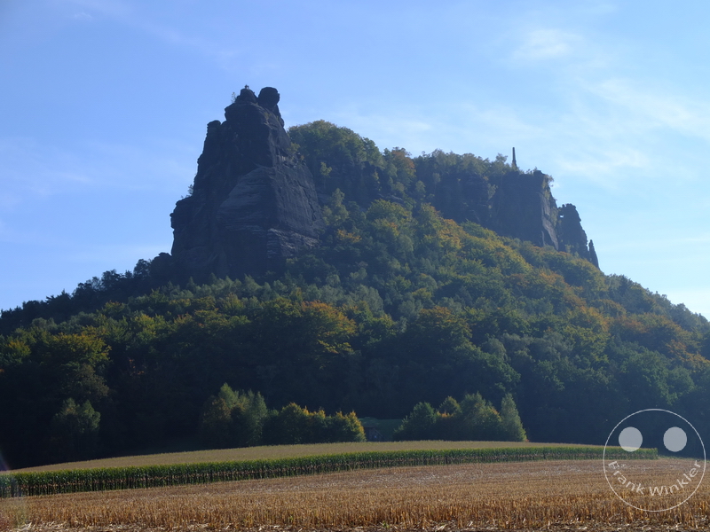 Deutschland - Sächsische Schweiz - Lilienstein - Bewaldeter Hügel mit Felsformation, abgeerntetes Feld, klarer blauer Himmel.