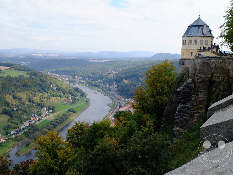 Deutschland - Festung Königstein - Großer Felsen mit gelbem Gebäude und Plattform, Fluss schlängelt sich durch grüne Landschaft.