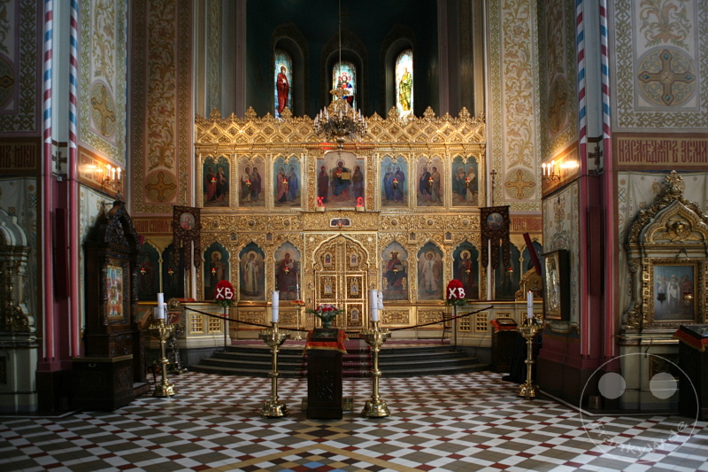 Estland - Tallinn - Ein beeindruckender Innenraum einer orthodoxen Kirche zeigt einen reich verzierten Ikonostase aus goldfarbenem Holz mit mehreren Ikonen und religiösen Bildern, flankiert von Kerzenständern und sorgfältig gestalteten Dekorationen. Der Boden ist mit einem geometrischen Fliesenmuster gestaltet. Im Hintergrund sind bunte Kirchenfenster zu sehen.