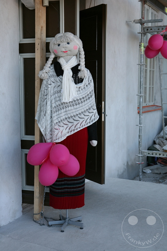 Estland - Tallinn - Stoffpuppe mit Zöpfen und Schal hält rosafarbene Luftballons vor einem Gebäude auf Ständer.