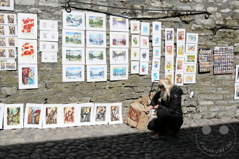 Estland - Tallinn - Kunstwerke an Steinwand, Person kniet und betrachtet Gemälde, große Tasche auf Kopfsteinpflaster.