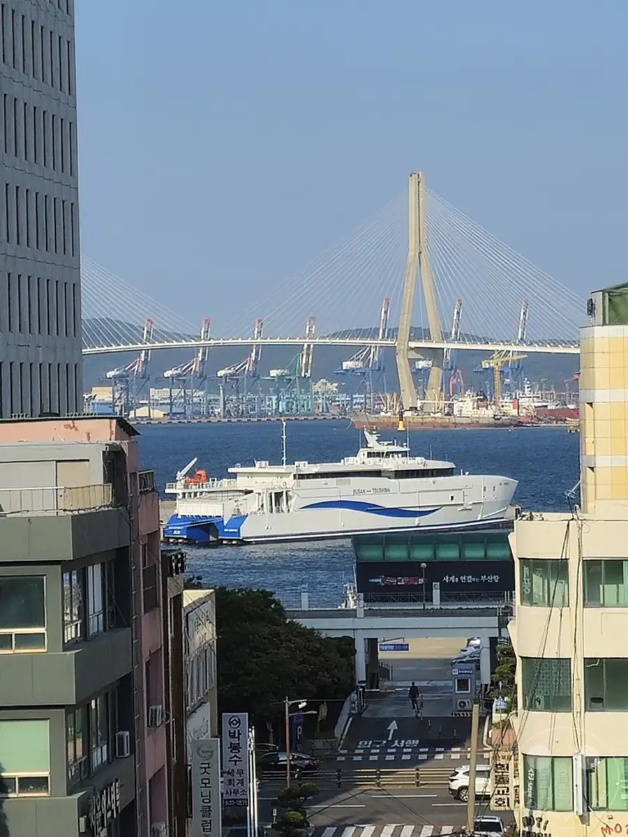 Eine große Fähre mit dem Namen 'Busan - Tsushima' fährt auf dem ruhigen Wasser des Hafens von Busan in Südkorea. Im Hintergrund erhebt sich majestätisch die Busan Harbor Bridge, ein beeindruckendes Wahrzeichen, das die moderne Infrastruktur der Stadt symbolisiert. Die Szene vermittelt ein Gefühl von urbaner Lebendigkeit und maritimer Bedeutung unter einem klaren blauen Himmel.