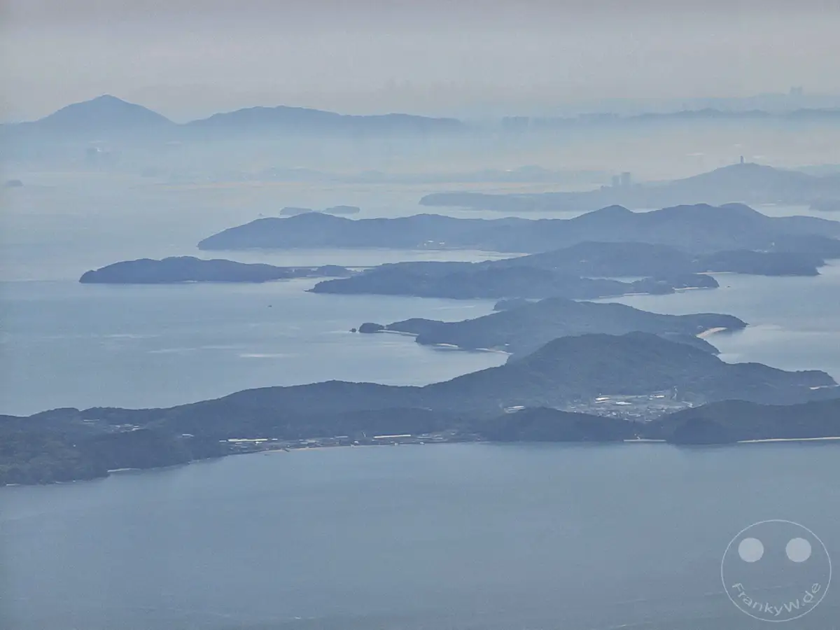 Ein malerischer Blick auf eine Reihe von dicht bewachsenen Inseln, die sich aus dem ruhigen Meer erheben, während ein leichter Nebel die fernen Umrisse einer Stadt und Berge umhüllt. Die Szenerie vermittelt eine friedliche und weite Atmosphäre während des Anflugs. Die grünen Inseln und das weite Wasser schaffen eine beruhigende Landschaft.