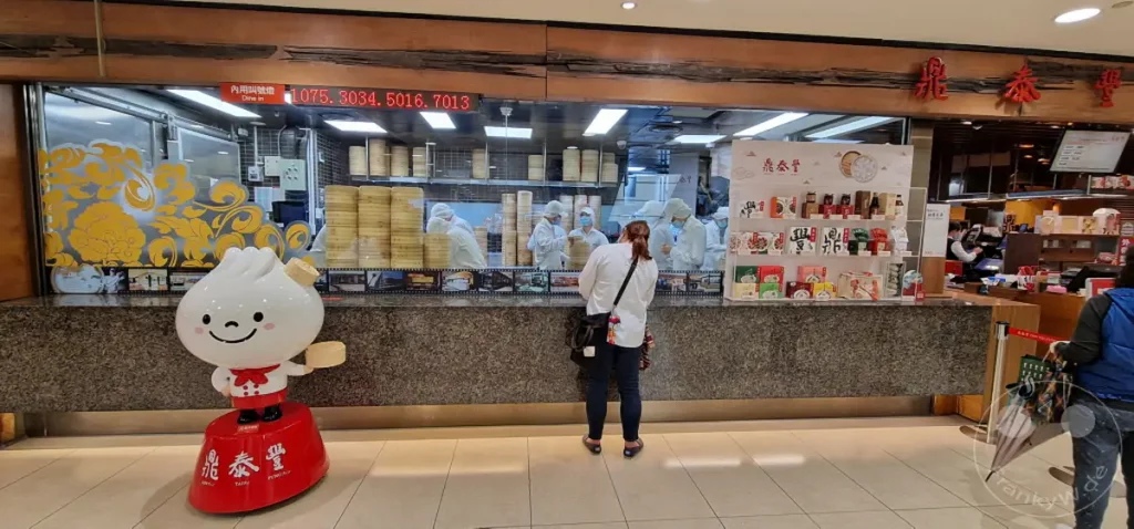 In einem Lebensmittelbereich des Sogo Kaufhauses in Taipeh stehen Kunden an einem Schalter, um authentisches Dim Sum zu bestellen. Hinter der Theke bereiten Mitarbeiter in Schutzbekleidung die gedämpften Köstlichkeiten in Bambusbehältern vor. Die Szene vermittelt eine geschäftige und authentische Atmosphäre für asiatische Speisen und Einkäufe.