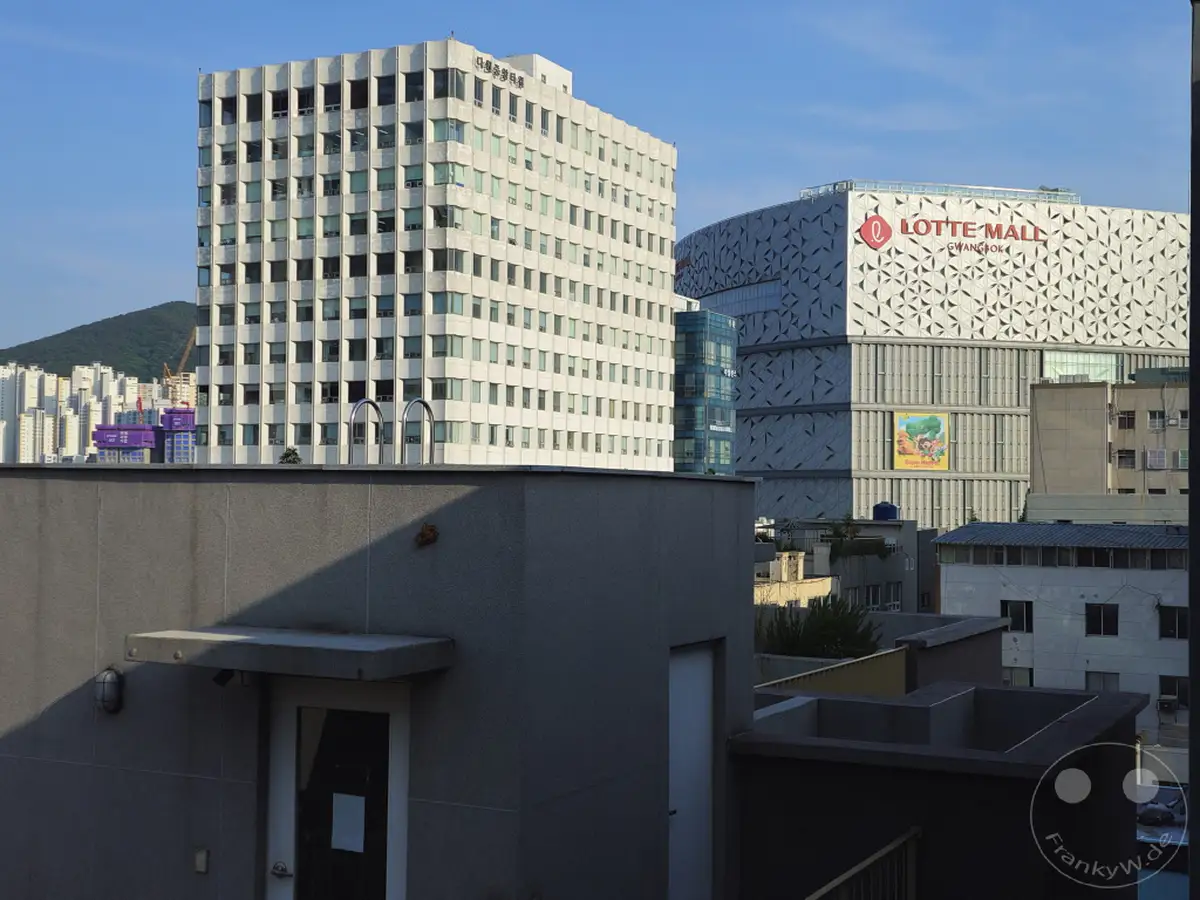 Die Lotte Mall in Busan, Südkorea, dominiert die urbane Skyline neben weiteren modernen Bürogebäuden. Die klare blaue Himmelskulisse betont die architektonische Vielfalt der Stadt. Eine typische südkoreanische Stadtlandschaft unter tags.