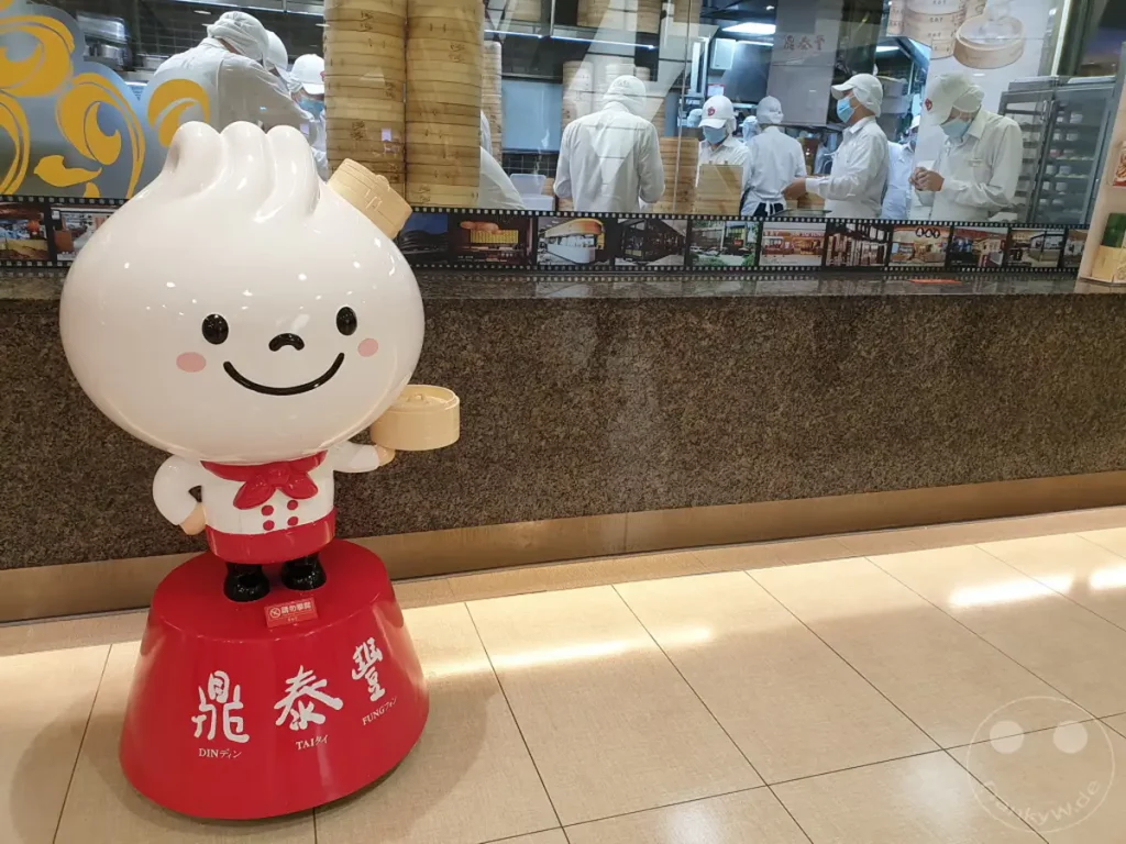 Ein niedliches Maskottchen in Form eines gedämpften Teigtäschchens steht vor einem Restaurant in Taipeh, Taiwan. Hinter dem Schalter sind Mitarbeiter zu sehen, die in dem bekannten Restaurant Din Tai Fung arbeiten, welches für seine Xiao Long Bao berühmt ist. Die Szene vermittelt eine fröhliche und kulinarische Atmosphäre.
