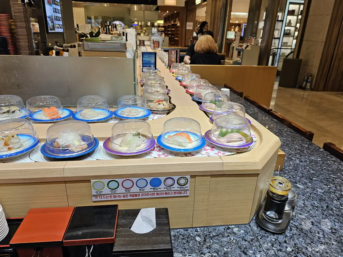 Eine breite Auswahl an Sushi-Gerichten wird auf einem Laufband in einem Restaurant in Südkorea präsentiert. Die kleinen Teller, jedes mit einer durchsichtigen Haube versehen, sind appetitlich aufgereiht und laden zum Probieren ein. Das helle und saubere Ambiente des Lotte Mall food court verspricht ein angenehmes kulinarisches Erlebnis in Busan.