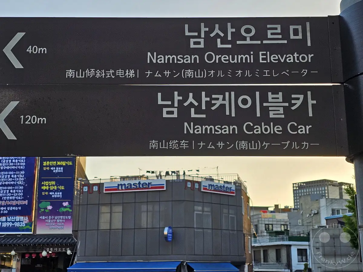 Eine Wegweiser-Beschilderung in Seoul, Südkorea, weist den Weg zur Namsan Oreumi Elevator und zur Namsan Cable Car. Im Hintergrund sind typische Stadtgebäude und der Verkehr in der Nähe des Namsan-Berges zu sehen. Die Beschilderung bietet sowohl koreanische als auch englische und chinesische Schriftzeichen für die Orientierung der Besucher.