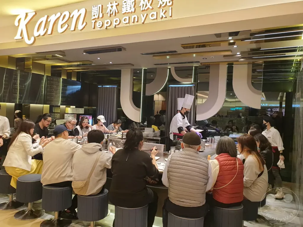 Gäste sitzen an einer Theke in einem Teppanyaki-Restaurant im Sogo Kaufhaus in Taipeh, Taiwan, und beobachten die Zubereitung ihrer Speisen. Ein Koch in Kochmütze bereitet die Gerichte vor den Augen der Anwesenden zu. Die Atmosphäre ist belebt, während die Gäste ihren kulinarischen Abend genießen.