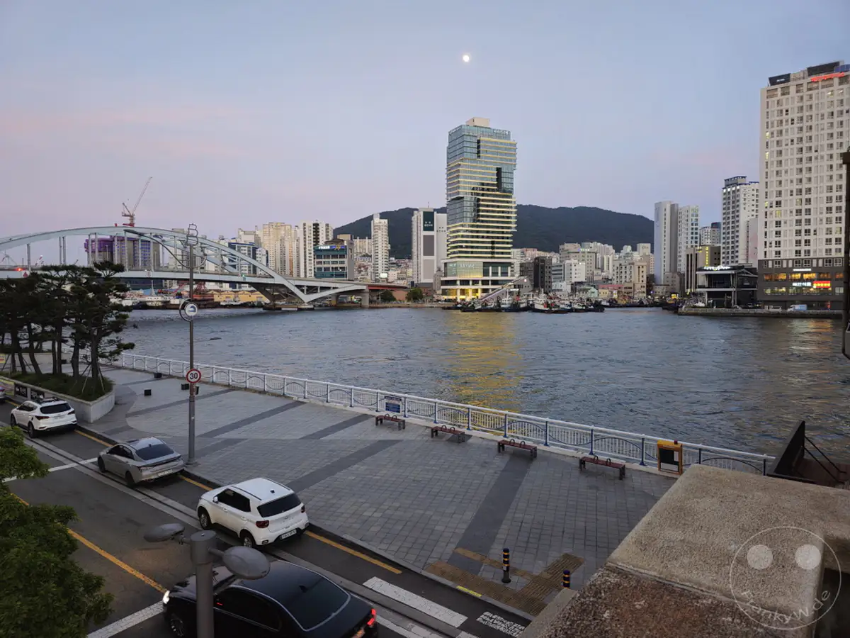 Die Skyline von Busan in Südkorea erstreckt sich entlang des Hafens, beleuchtet von der Dämmerungsbeleuchtung. Die imposante Busan Bridge überspannt das Wasser und verbindet die städtischen Gebiete, während Boote im Hafen ruhen. Eine friedliche Atmosphäre liegt über der Stadt, die im sanften Licht des schwindenden Tages erstrahlt.