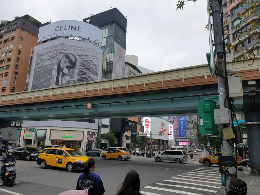 Eine belebte Straßenszene in Taipeh, Taiwan, mit der bekannten Sogo-Kaufhausfassade und Reklametafeln. Gelbe Taxis und Autos bewegen sich auf der mehrspurigen Straße unter einer erhöhten Brücke. Die Atmosphäre ist lebhaft und typisch für eine asiatische Metropole.