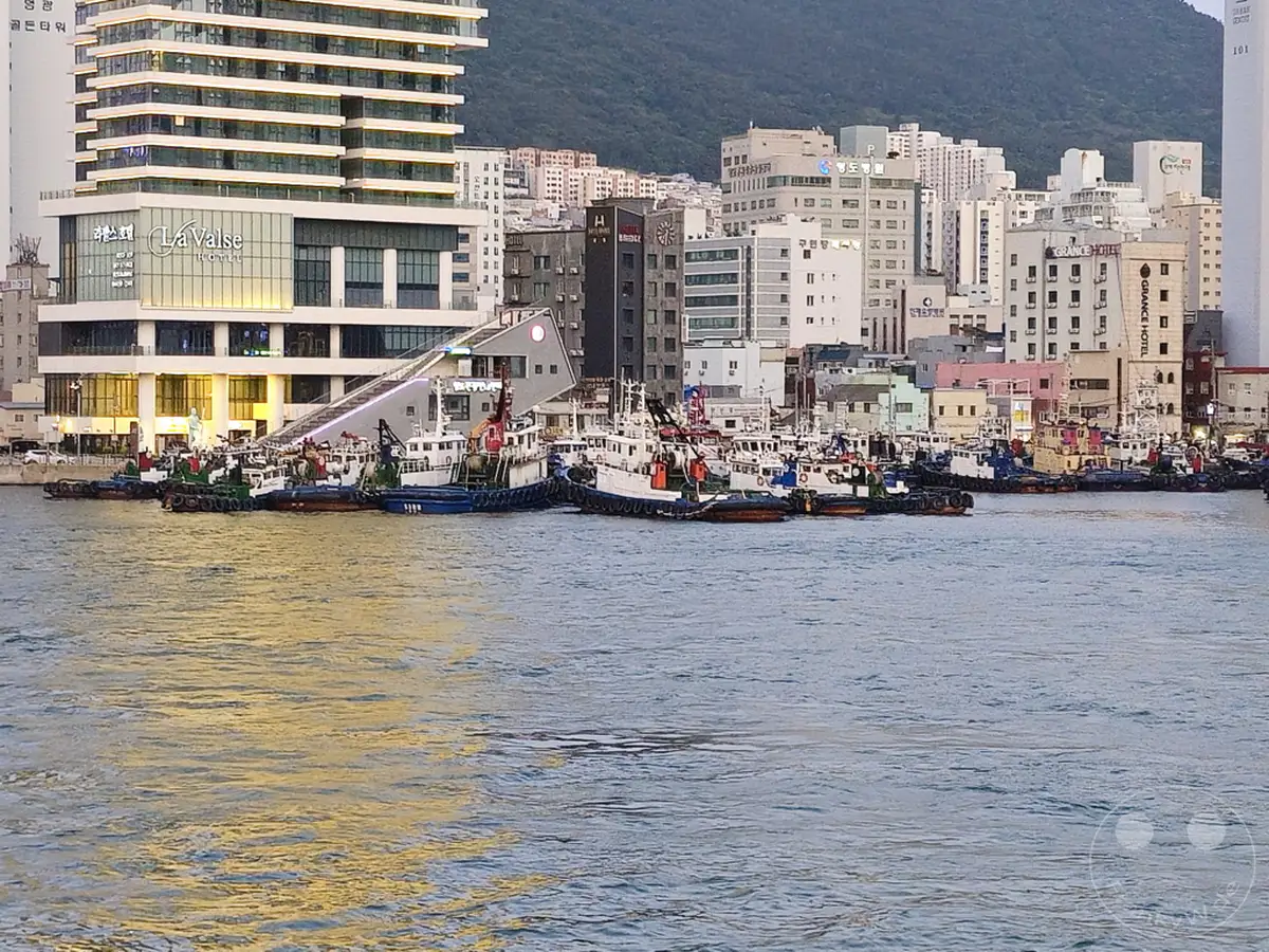Eine Reihe von Schleppern und kleineren Booten liegt im Hafen von Busan, Südkorea. Im Hintergrund erheben sich moderne Gebäude und ein Hügel, der mit Vegetation bedeckt ist. Das ruhige Wasser spiegelt das sanfte Licht des späten Tages und schafft eine friedliche Atmosphäre im belebten Hafen.