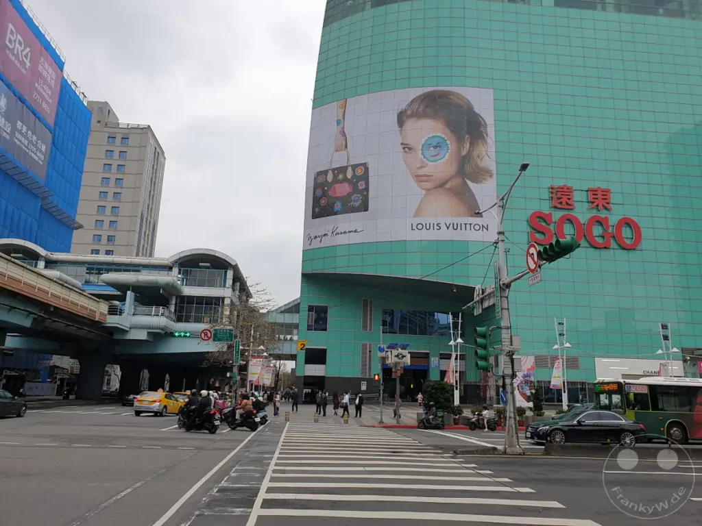 In Taipeh, Taiwan, markiert das Sogo Kaufhaus mit seiner charakteristischen grünen Fassade und einer großen Werbetafel ein zentrales Element der städtischen Landschaft. Der belebte Verkehr mit Autos, Motorrollern und einer Bushaltestelle unterstreicht die Lebendigkeit der Metropole. Der Himmel ist bedeckt und verleiht der Szene eine gedämpfte, aber geschäftige Atmosphäre.