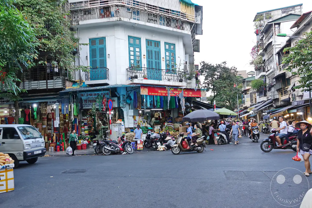 Das geschäftige Treiben im Old Quarter von Hanoi, Vietnam, ist geprägt von Motorrollern und Ständen, die Waren feilbieten. Die Szenerie wird von traditionellen Häusern mit bunten Fassaden und Balkonen untermalt, während Menschen ihren täglichen Geschäften nachgehen. Die Atmosphäre ist lebendig und authentisch, typisch für eine südostasiatische Metropole.