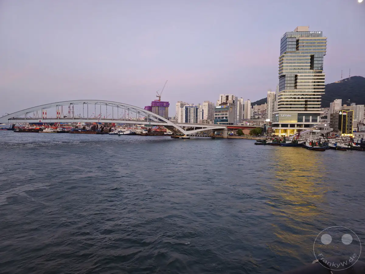 Die beleuchtete Busan Bridge überspannt einen belebten Hafen in Südkorea bei Dämmerung. Moderne Hochhäuser, darunter ein markantes glasverkleidetes Gebäude, spiegeln sich im Wasser, während kleine Boote am Ufer liegen. Die Szene vermittelt eine ruhige, urbane Atmosphäre, die das geschäftige Treiben der Stadt einfängt.