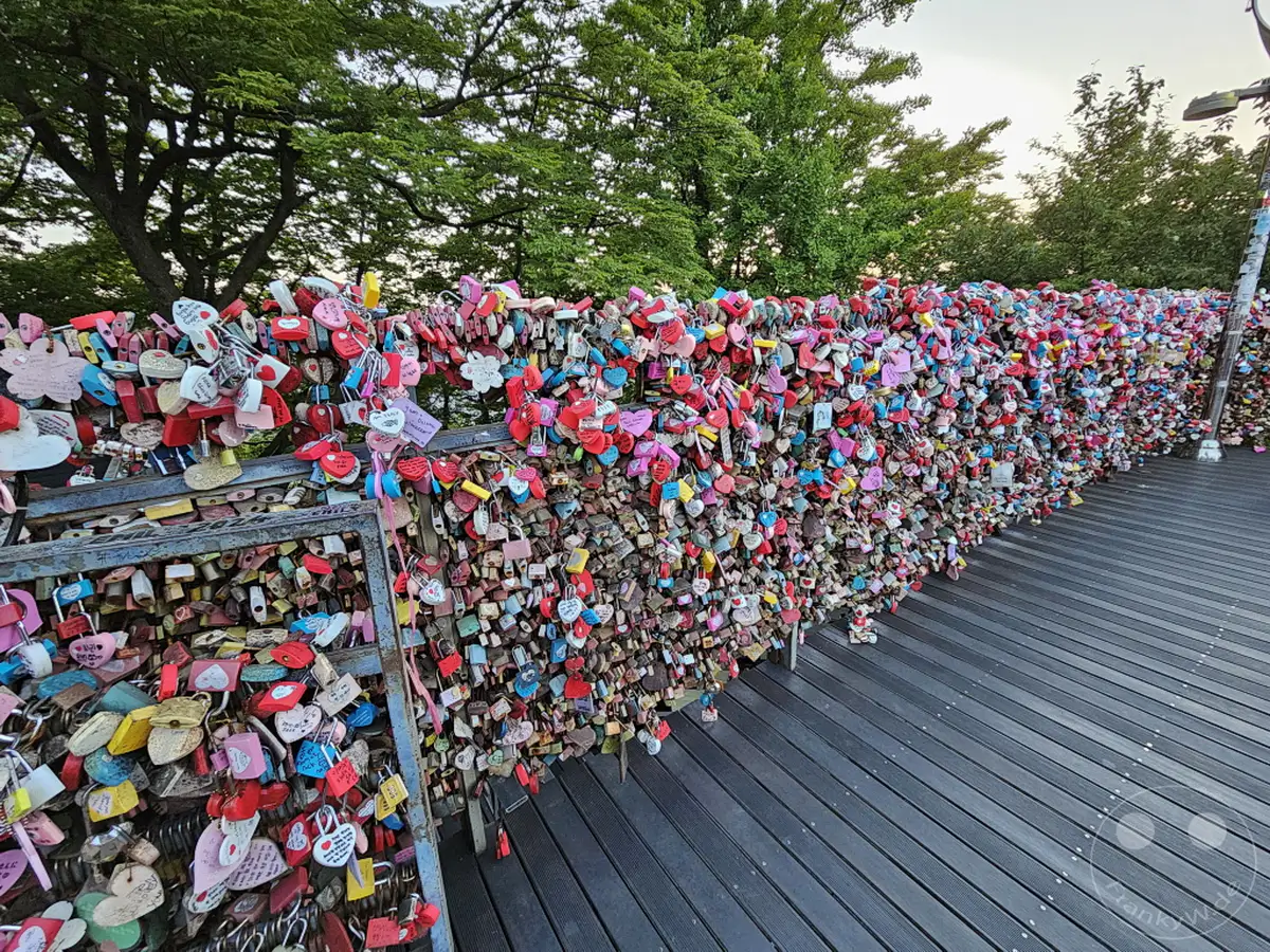 Eine überwältigende Wand aus tausenden bunten Liebesschlössern ziert die Brüstung der Aussichtsplattform des Namsan Tower in Seoul, Südkorea. Jedes Schloss, oft mit Namen und Daten versehen, symbolisiert die unzähligen Liebesbekundungen und Hoffnungen von Besuchern. Die dichte Anordnung der Schlösser schafft ein lebendiges und bewegendes Mosaik vor der Kulisse des grünen Baumbestands.