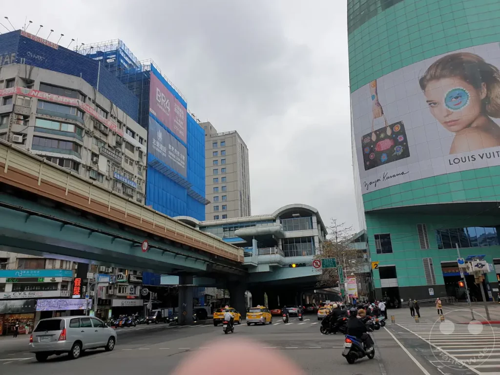 Die Aufnahme vermittelt eine geschäftige Straßenszene in Taipeh, Taiwan, mit einem modernen Gebäude, das eine Louis Vuitton-Werbung mit Yayoi Kusama-Kunst ziert. Der Verkehr fließt unter einer Hochbahn, während Menschen und Fahrzeuge die belebte urbane Landschaft beleben. Das Wetter ist bedeckt, was der Szene eine gedämpfte, alltägliche Atmosphäre verleiht.