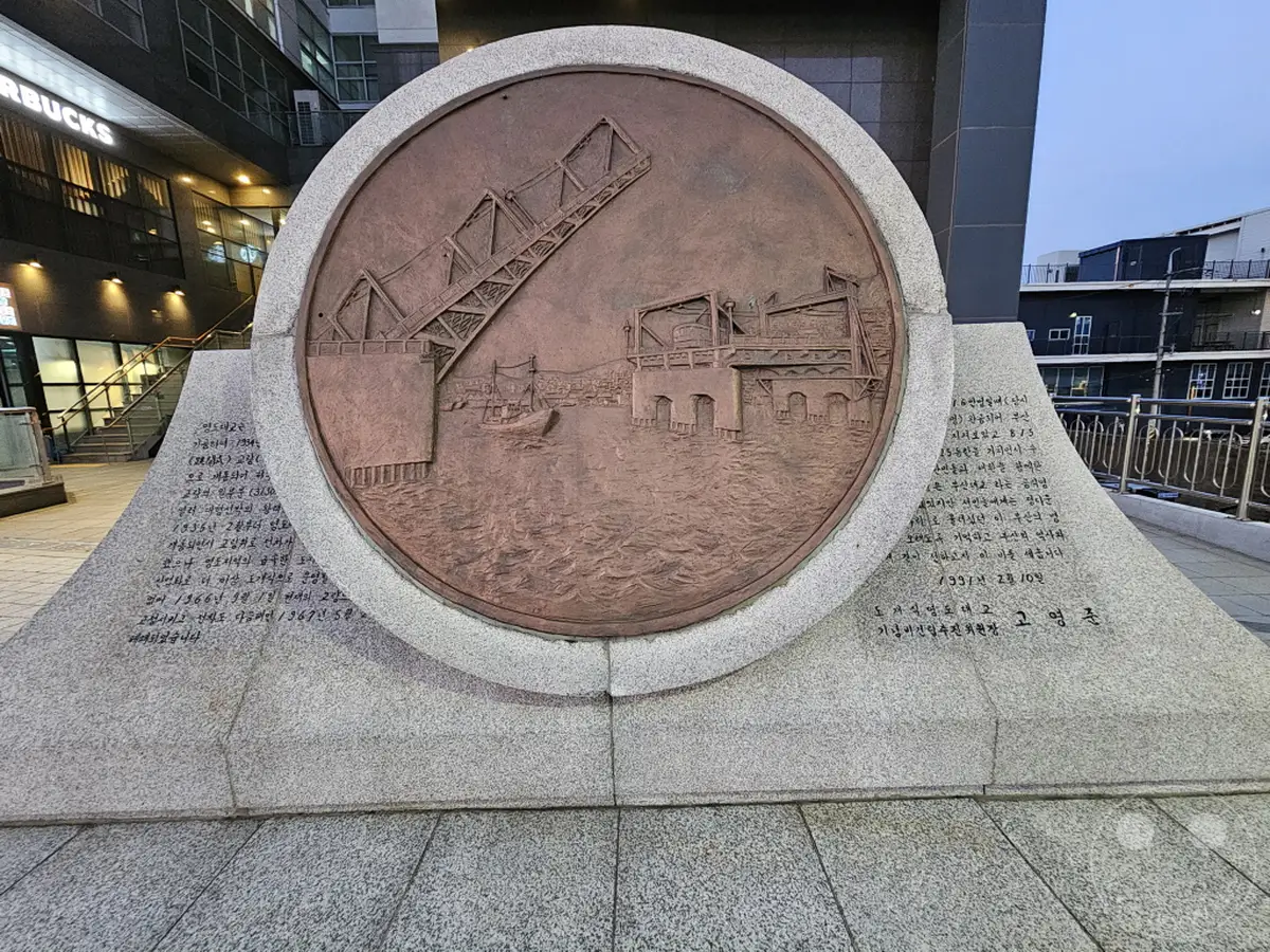 Ein bronzenes Relief auf einem Steinsockel ehrt die Yeongdo-Brücke in Busan, Südkorea. Das Kunstwerk zeigt die historische Bedeutung der Brücke mit einer Schiffsdarstellung und maritimen Elementen. Die Inschriften auf dem Sockel geben Einblicke in die Geschichte und die Bedeutung des Denkmals.