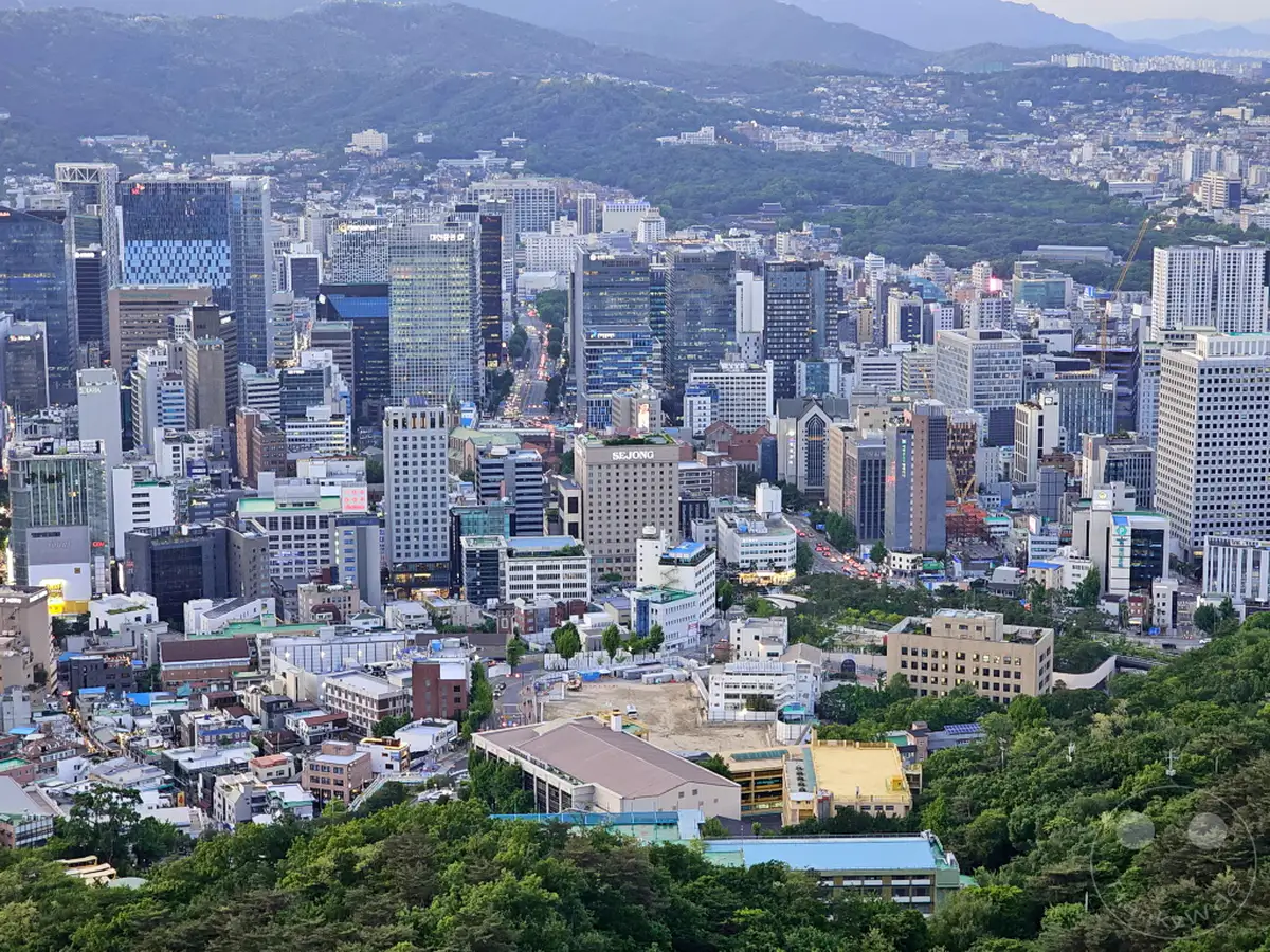 Ein weites Panorama von Seoul, Südkorea, fängt die beeindruckende Dichte urbaner Architektur gegen die sanften Hügel der umliegenden Berge ein. Die Abenddämmerung taucht die Stadt in ein mildes Licht, wobei die schimmernden Fenster der Wolkenkratzer und die geschäftigen Straßen ein Gefühl von Dynamik und Erhabenheit vermitteln. Diese Aussicht vom N Seoul Tower bietet einen atemberaubenden Einblick in das moderne Leben und die natürliche Schönheit, die Seoul prägen.