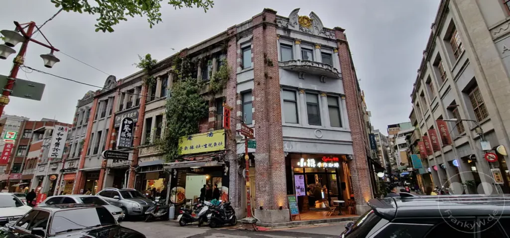Historische Gebäude säumen die belebte Dihua Street in Taipeh, Taiwan, unter einem bewölkten Himmel. Die Fassaden der Geschäfte und Wohnhäuser zeugen von einer vergangenen Ära, während das geschäftige Treiben auf der Straße eine lebendige Atmosphäre schafft. Die Architektur mit ihren traditionellen Elementen und dem teils überwucherten Zustand verleiht der Szene einen einzigartigen Charme.