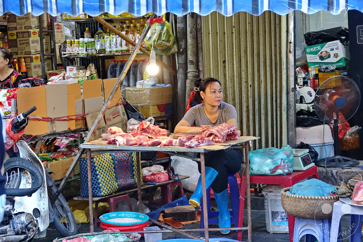 In Hanoi, Vietnam, spielt sich das geschäftige Treiben im Old Quarter – die Altstadt ab, wo eine Fleischverkäuferin an einem Stand sitzt. Rohes Fleisch liegt ausgebreitet, während die Händlerin den Markt beobachtet und auf Kunden wartet. Die Szene strahlt die authentische Atmosphäre eines lebhaften vietnamesischen Marktes aus.