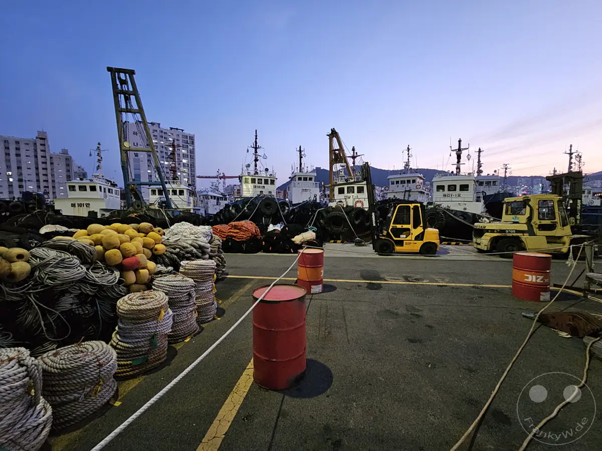 In Südkorea, am Busan Port, liegen am frühen Morgen Fischerboote und Kräne bereit. Angehäuftes Tauwerk und leere Fässer säumen den Kai, während die aufgehende Sonne den Himmel in sanften Farben erhellt. Eine ruhige, aber geschäftige Atmosphäre liegt über dem Hafen vor dem Beginn des Tages.