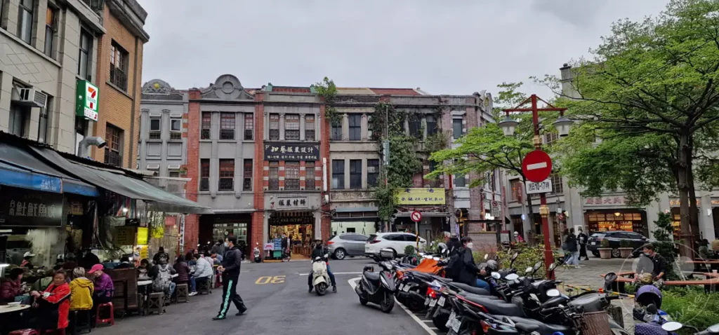 Die Dihua Street in Taipeh, Taiwan, präsentiert sich als belebte Marktstraße mit traditionellen Gebäuden und vielen geparkten Mopeds. Die Szene vermittelt das geschäftige Treiben eines asiatischen Marktes, während Menschen an Imbissständen und Geschäften verweilen. Eine Mischung aus Altem und Neuem prägt das Bild, eingefangen an einem bewölkten Tag.