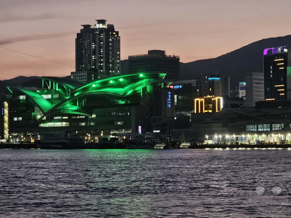 Die beleuchtete Skyline von Busan mit dem Busan Port bei Einbruch der Dunkelheit, erleuchtet von den grünen Lichtern eines modernen Gebäudes. Boote liegen im ruhigen Wasser vor der Küstenfront, während die Silhouette der Berge im Hintergrund gegen den dämmrigen Himmel aufragt. Die Szene strahlt eine lebendige, aber friedliche Atmosphäre aus.