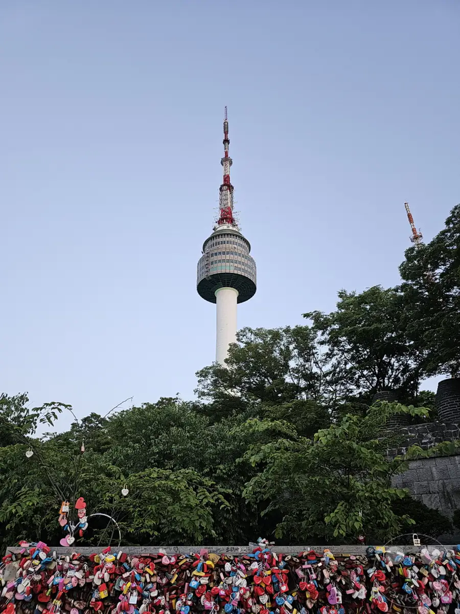 Der Namsan Tower in Seoul, Südkorea, ragt majestätisch in den klaren Himmel. An den Geländern sind unzählige Liebesschlösser in allen Farben befestigt, ein Symbol für die ewige Liebe vieler Paare. Üppige grüne Bäume umgeben die Basis des Turms und vervollständigen das idyllische Bild dieser berühmten Touristenattraktion.