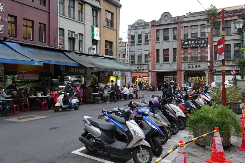 Eine belebte Straße in Taipeh, Taiwan, an einem bewölkten Tag. Roller parken entlang der Dihua Street, während Menschen in den vielen Geschäften und Restaurants einkaufen und essen. Die Szene vermittelt einen lebendigen Eindruck des täglichen Lebens in dieser historischen Marktstraße.