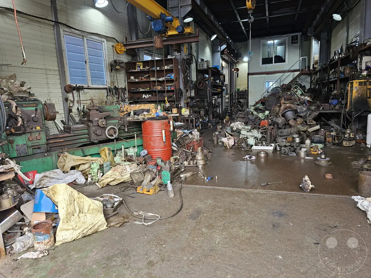 Eine überladene Werkstatt im Hafen von Busan, Südkorea, ist gefüllt mit alten Maschinen, Ersatzteilen und Geräten. Der Boden ist nass und schmutzig, was auf regen Betrieb oder mangelnde Instandhaltung hindeutet. Die Szene vermittelt einen Eindruck von industrieller Hektik und dem Verfall über die Zeit.