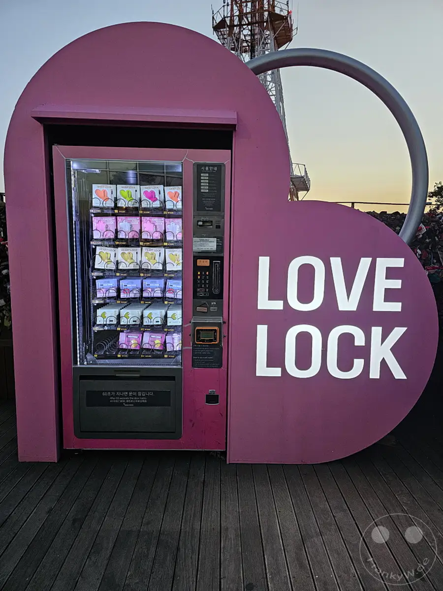 Ein pinker Verkaufsautomat, der als Herz gestaltet ist, bietet auf dem Namsan Tower in Seoul, Südkorea, "Love Locks" zum Kauf an. Die Verkaufsstelle ist Teil des beliebten "Love Lock"-Erlebnisses, bei dem Paare Vorhängeschlösser anbringen, um ihre Liebe zu symbolisieren. Die Atmosphäre ist romantisch und die Abenddämmerung taucht die Szene in ein sanftes Licht.