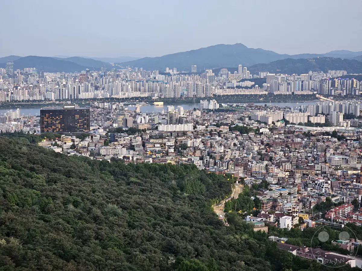 Die beleuchtete Skyline von Seoul erstreckt sich bis zu den fernen Bergen, während die Sonne untergeht und den Himmel in sanfte Pastelltöne taucht. Von der Anhöhe des N Seoul Towers bietet sich ein atemberaubender Panoramablick über die geschäftige Metropole Südkoreas. Der Kontrast zwischen den dichten städtischen Gebieten und der umgebenden Natur schafft eine friedliche und zugleich dynamische Atmosphäre.