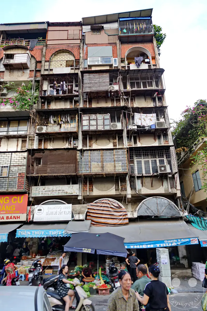Die engen Gassen des Old Quarter in Hanoi, Vietnam, sind belebt von Marktständen und Passanten. Das tägliche Leben entfaltet sich vor einer Kulisse aus dicht gedrängten, traditionellen Wohngebäuden mit sichtbaren Einflüssen aus verschiedenen Bauperioden. Die Szene vermittelt einen authentischen Eindruck des pulsierenden Stadtlebens.
