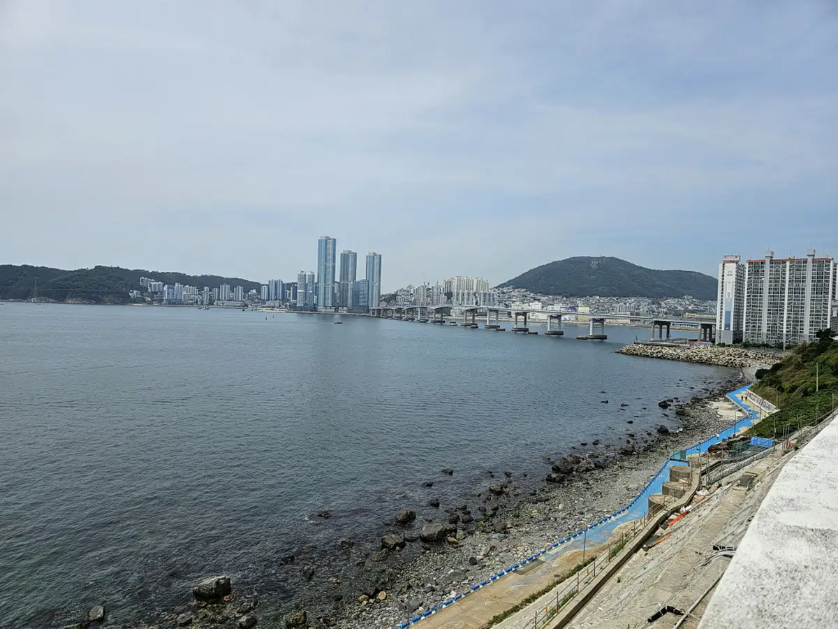 Die weite See erstreckt sich vor der Küste von Busan, Südkorea. Am Ufer grenzt das Huinnyeoul Culture Village an einen steinigen Strand und eine malerische Promenade, die sich entlang der felsigen Küste schlängelt. Im Hintergrund erhebt sich eine beeindruckende Stadtkulisse mit modernen Hochhäusern, überragt von einer markanten Bergformation und einer langen Brücke, die die Bucht überspannt.