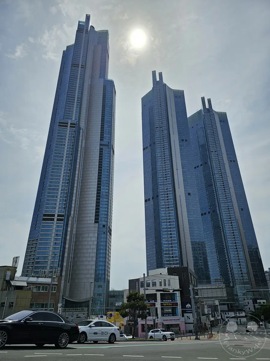 Der Himmel über Busan, Südkorea, ist an diesem sonnigen Tag von mehreren beeindruckenden Wolkenkratzern dominiert. Die gläsernen Fassaden der Hochhäuser spiegeln das gleißende Sonnenlicht wider und erzeugen eine moderne, architektonische Stadtansicht. Autos bewegen sich auf einer breiten Straße im Vordergrund, während die imposanten Gebäude in den Himmel ragen und einen Eindruck von Fortschritt und Urbanität vermitteln.