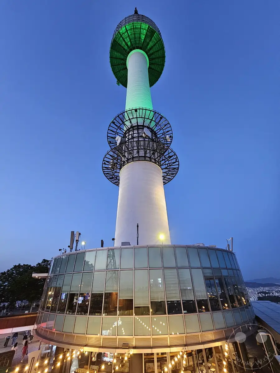 Der N Seoul Tower ragt in der Abenddämmerung empor, sein oberer Teil ist in ein lebendiges Grün getaucht. Ein runder Glasanbau mit Beleuchtung und Lichterketten umgibt die Basis des Turms und reflektiert die Lichter der Stadt. Die klare blaue Abendhimmel bildet einen eindrucksvollen Kontrast zur beleuchteten Struktur und schafft eine magische Atmosphäre über Seoul.