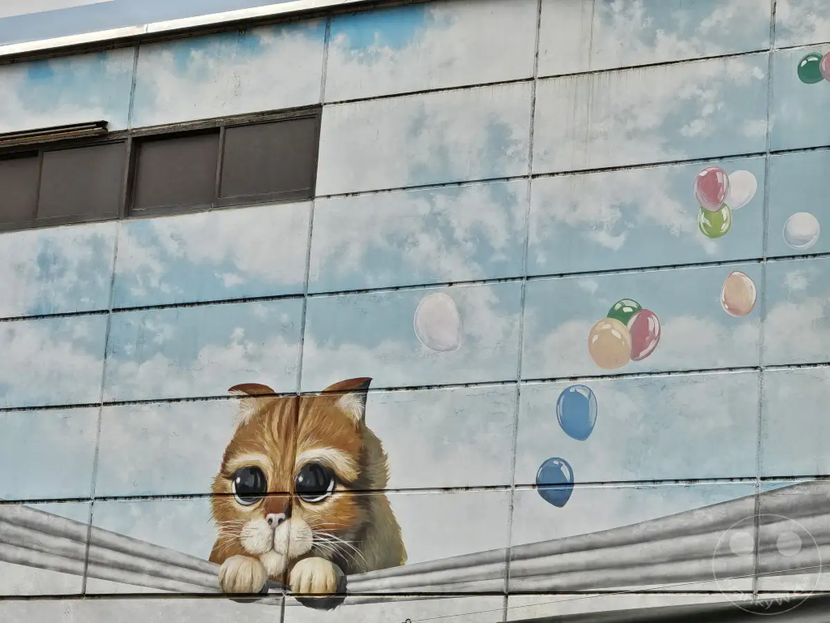 Ein lebhaftes Graffiti auf einer Hauswand in Seoul, Südkorea, präsentiert eine süße Katze mit großen Augen, die hinter einer Stoffbahn hervorlugt. Mehrere bunte Ballons steigen in den gemalten Himmel auf und erzeugen eine verspielte und fantasievolle Atmosphäre. Dieses Kunstwerk belebt die Einkaufsstraße und zieht die Blicke von Passanten auf sich.