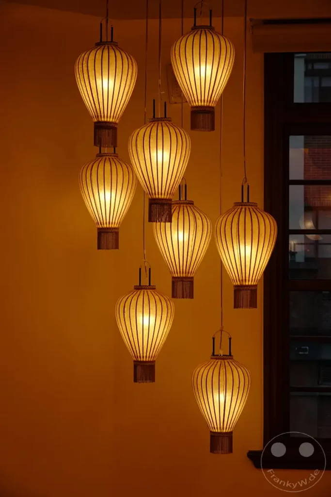 Eine Gruppe von asiatisch inspirierten Lampions, beleuchtet mit warmem Licht, hängt von der Decke in einem Innenraum. Die traditionellen Lampions aus Papier und Holz zieren den Raum und erzeugen eine gemütliche und einladende Atmosphäre, typisch für die Dihua Street in Taipeh, Taiwan. Das gedämpfte Licht und die symmetrische Anordnung der Laternen schaffen ein harmonisches Gesamtbild.