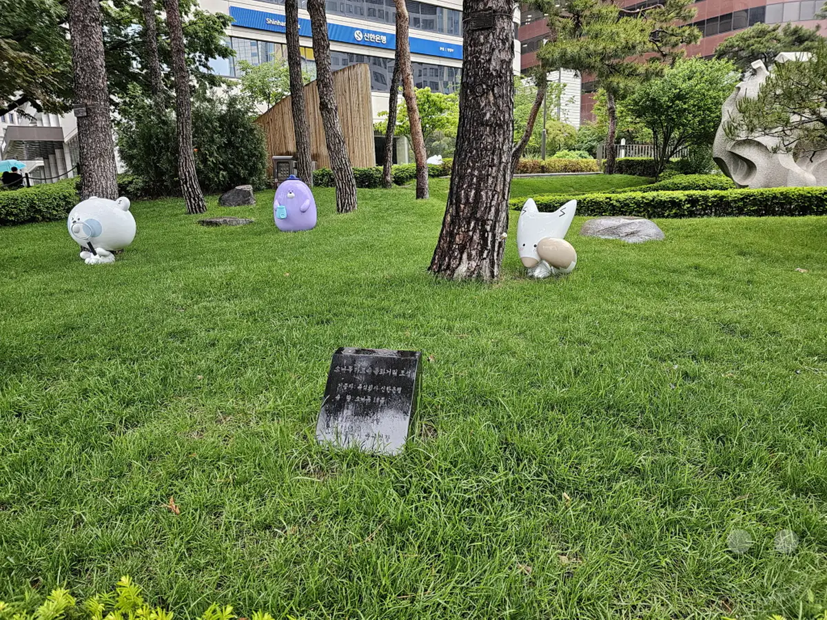 Eine idyllische Grünanlage im Herzen von Seoul, Südkorea, beherbergt mehrere verspielte Skulpturen, darunter eine weiße runde Figur mit Brille und eine lila Kreatur mit Hut. Im Hintergrund erhebt sich das moderne Gebäude der Shinhan Bank, das einen Kontrast zur natürlichen Umgebung bildet. Der sorgfältig gepflegte Rasen und die Bäume schaffen eine ruhige und einladende Atmosphäre inmitten des städtischen Trubels.