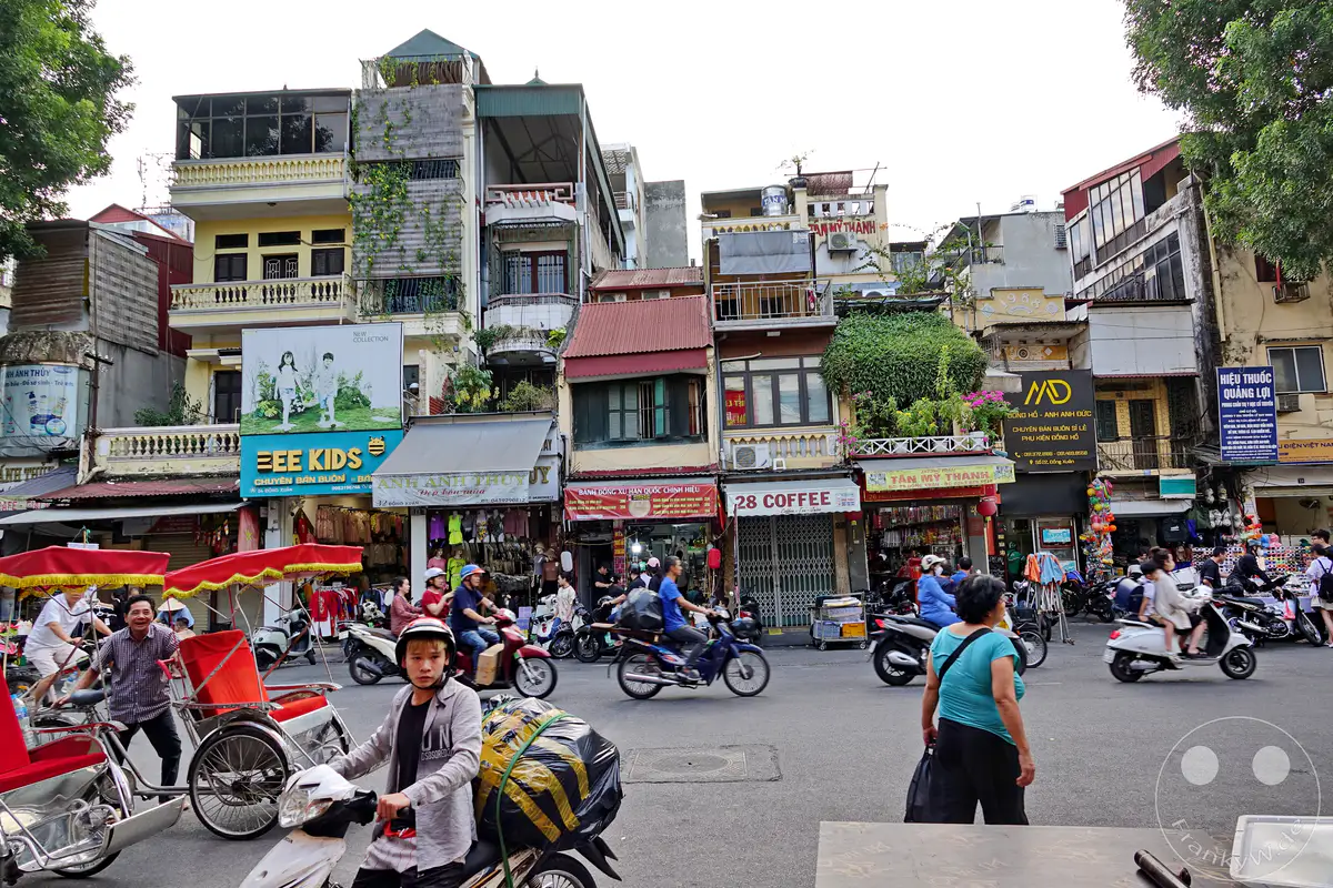 Im Old Quarter von Hanoi, Vietnam, herrscht geschäftiges Treiben auf den Straßen. Zahlreiche Geschäfte säumen die engen Gassen und bieten Waren aller Art an, während Motorroller und Rikschas den Verkehr beleben. Die Szene vermittelt einen lebendigen Eindruck des Alltags in dieser pulsierenden Stadt.