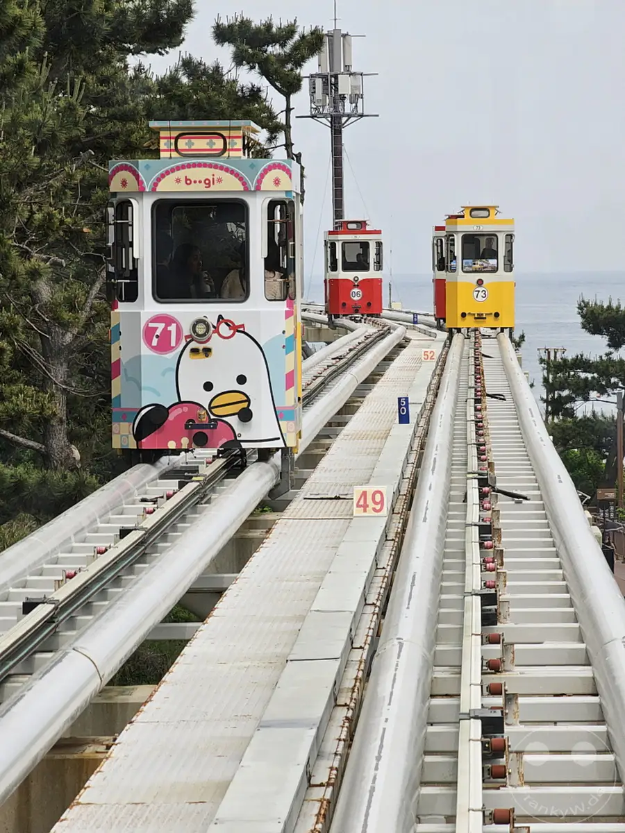 Die Küstenbahn im Haeunde Blueline Park in Busan, Südkorea, fährt auf ihrer Strecke entlang des Meeres. Mehrere bunte Züge, darunter der nummerierte "71" mit einer Pinguin-Dekoration, sind auf der erhöhten Bahn zu sehen. Die Szenerie vermittelt eine fröhliche und entspannte Urlaubsatmosphäre mit Blick auf das Meer und die Küstenlandschaft.