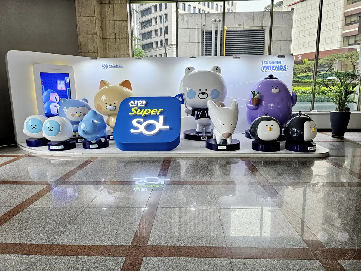 In Seoul, Südkorea, präsentiert sich die Shinhan Bank mit ihren beliebten Shinhan Friends Charakteren und der Super SOL Ausstellung. Die fröhlich gestalteten Figuren und das leuchtende "Super SOL" Logo schaffen eine einladende und moderne Atmosphäre. Die Szene spielt in einem hellen Foyer, wo die Charaktere auf Podesten ausgestellt sind, und spiegelt die digitale Ausrichtung des Unternehmens wider.