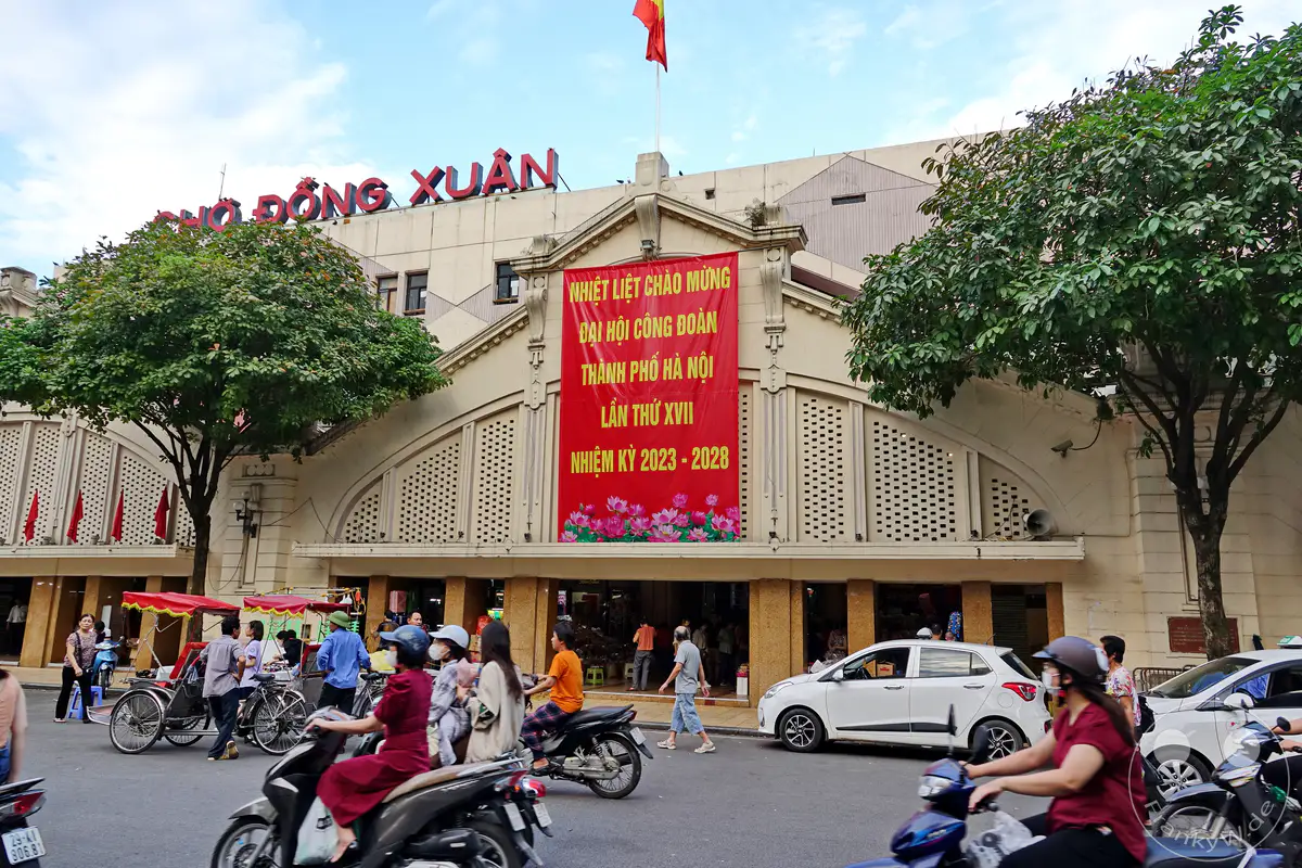 Der geschäftige Dong Xuan Markt im Herzen von Hanois Old Quarter ist ein Zentrum des städtischen Lebens. Motorroller und Autos bewegen sich durch die belebten Straßen vor der Marktfront, während bunte Banner die Fassade schmücken. Die Atmosphäre ist belebt und voller Energie, typisch für diese historische Stadtregion in Vietnam.