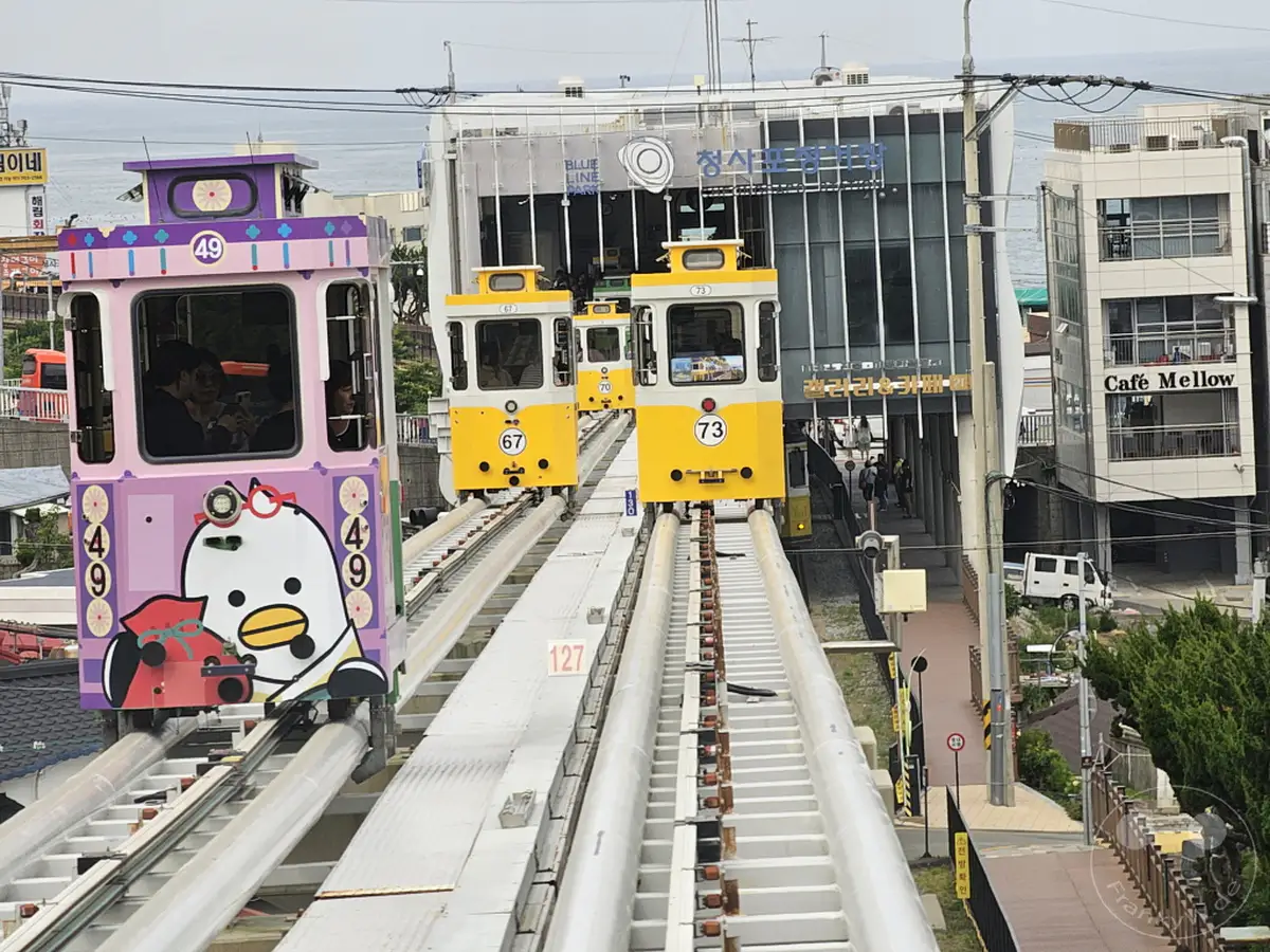 In Südkorea, Busan, verkehren farbenfrohe Züge auf der Küstenbahn im Haeundae Blueline Park. Die Züge, darunter ein lila Wagen mit einem Entenmotiv, fahren entlang der Gleise in der Nähe der Cheongsapo Station. Die Szene vermittelt eine fröhliche und touristische Atmosphäre entlang der Küste.