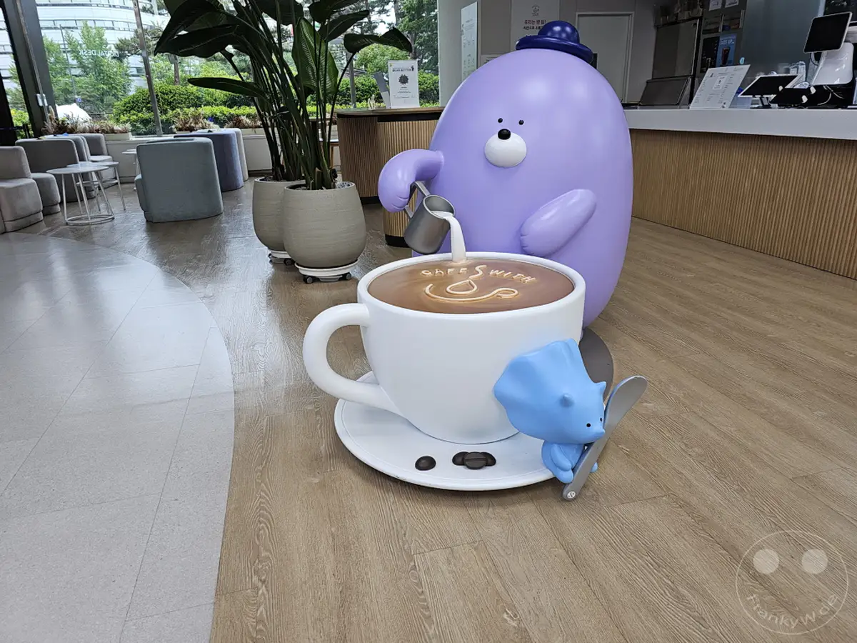 Eine interaktive Dekoration in einem Café-Bereich, vermutlich in der Nähe einer Shinhan Bank in Seoul, Südkorea, präsentiert eine riesige Kaffeetasse, aus der eine lila Figur gerade Getränk einschenkt. Neben der Tasse steht eine kleine blaue Figur, die einen Löffel hält, und schafft eine verspielte und einladende Atmosphäre. Der Boden und die umliegenden Möbel deuten auf ein modernes und stilvolles Interieur hin.