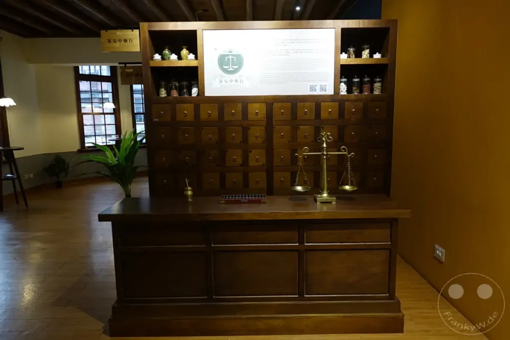 In Taipeh, Taiwan, an der Dihua Street, steht eine traditionelle Apotheke mit einem imposanten Holzschrank voller kleiner Schubladen, die einst Heilkräuter beherbergten. Daneben eine Waage, die an vergangene Zeiten und die sorgfältige Zubereitung von Arzneimitteln erinnert. Der warme Holzkasten und die gedämpfte Beleuchtung erzeugen eine Atmosphäre von Geschichte und Ruhe.