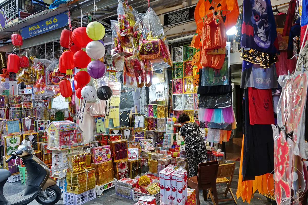 In Hanoi, Vietnam, präsentiert sich das Old Quarter – die Altstadt – mit einem lebhaften Straßenverkauf, der eine Fülle bunter Lampions und thematischer Kostüme anbietet. Die Szene ist gefüllt mit verschiedensten Artikeln, die von traditionellen Laternen bis hin zu Halloween-Verkleidungen reichen und eine festliche, kommerzielle Atmosphäre schaffen. Ein Rollerräder steht im Vordergrund, während im Hintergrund eine Verkäuferin die Waren arrangiert und die geschäftige Energie des Marktes unterstreicht.