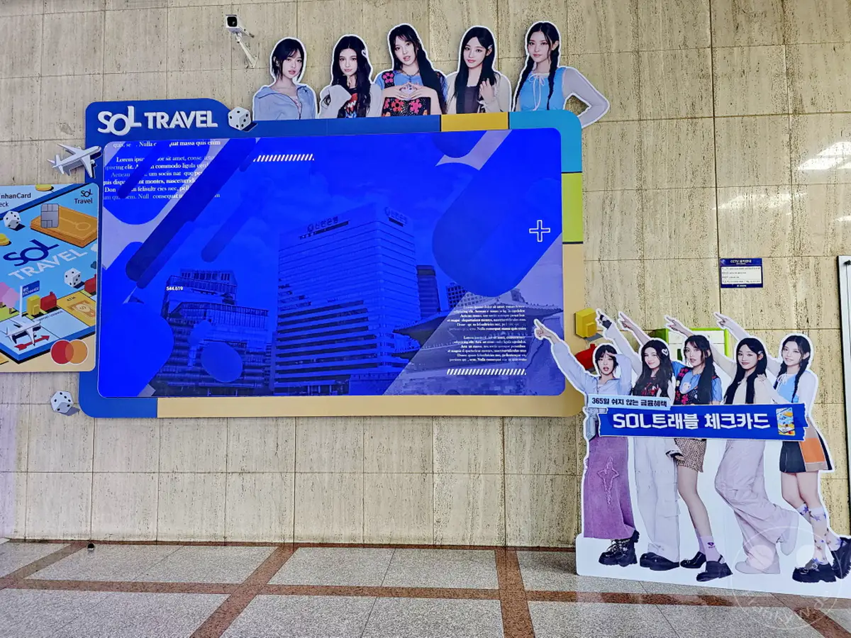 In Seoul, Südkorea, wirbt die Shinhan Bank für ihre SOL Travel Checkkarte mit einer auffälligen Installation. K-Pop-Idole posieren für die Kampagne und ziehen die Aufmerksamkeit der Passanten auf sich. Die bunte und moderne Darstellung vermittelt eine lebendige und ansprechende Atmosphäre für die Finanzdienstleistung.