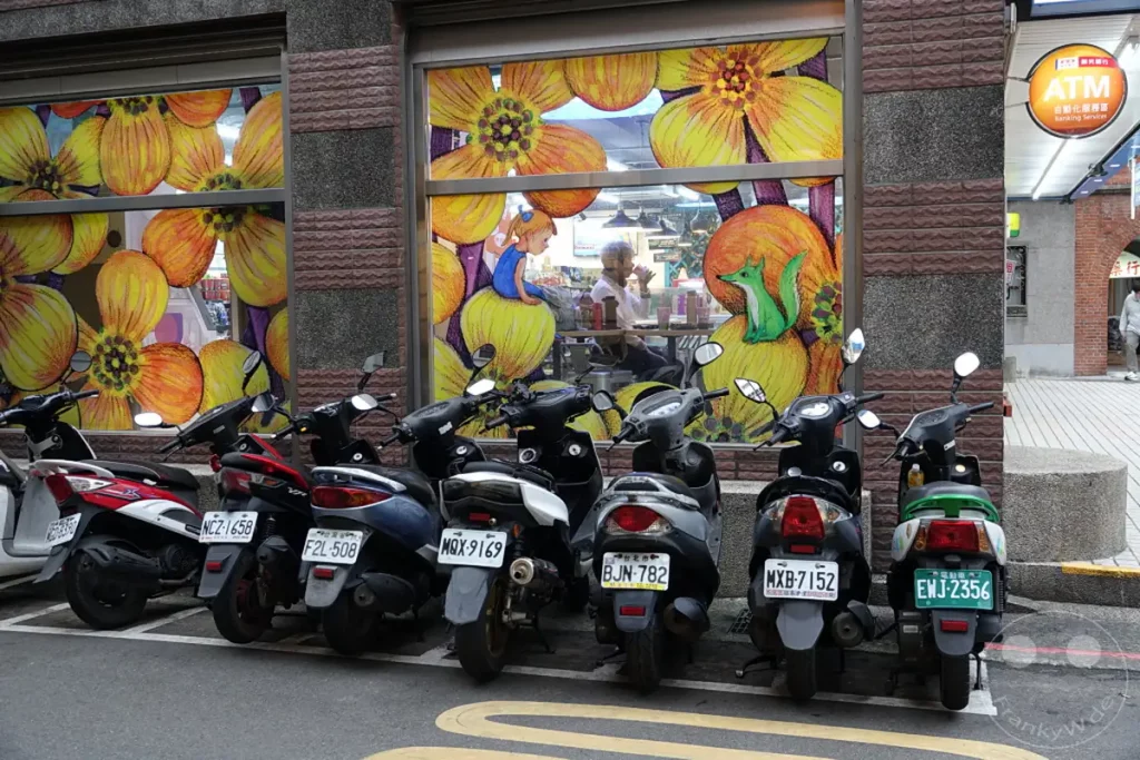 Mehrere Roller parken nebeneinander vor einer bunten Fassade in Taipeh, Taiwan. Die Fenster zeigen ein lebhaftes Blumenmuster mit gelben und orangen Blüten. Die Szene wirkt alltäglich und doch künstlerisch durch die farbenfrohe Wandgestaltung.